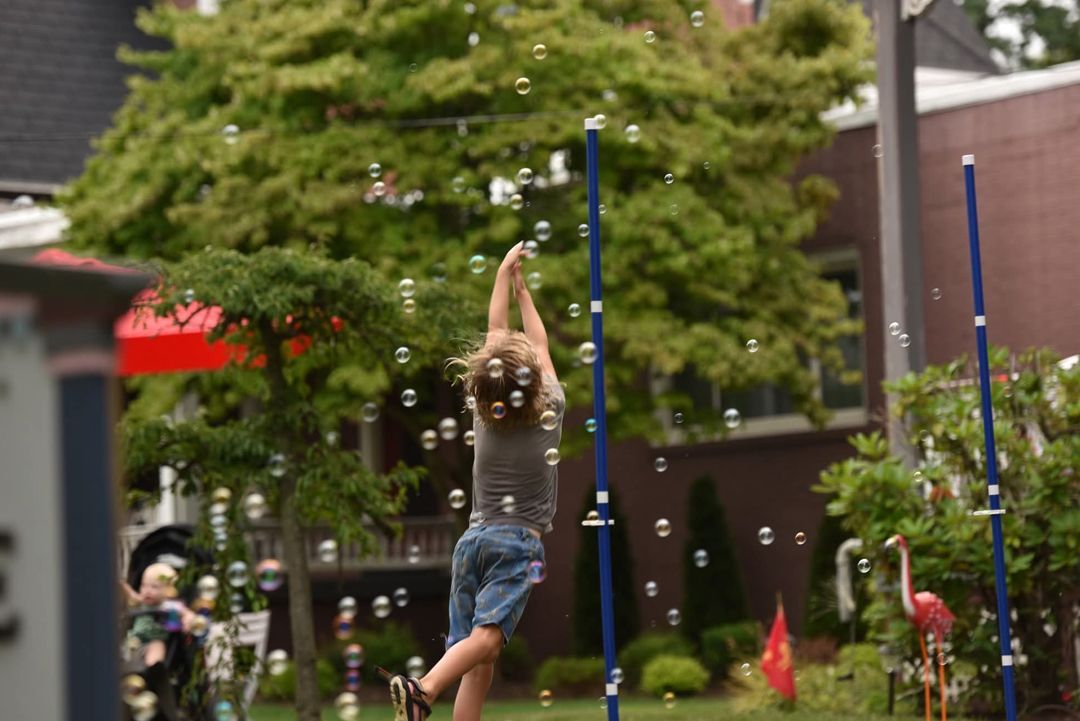 Child reaching for bubbles in a yard. They wear shorts and a grey top with a blurred out background of trees, buildings, and pink lawn flamingos.