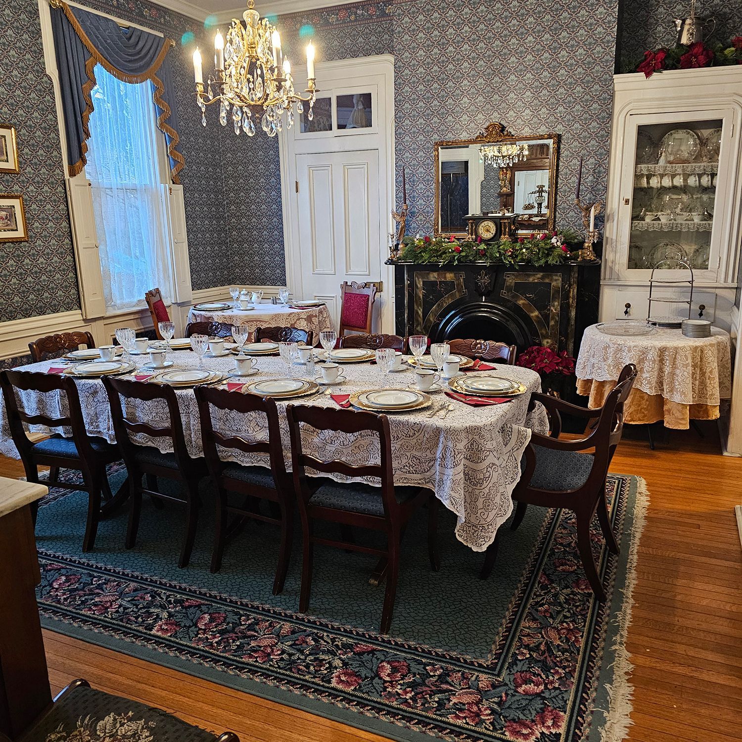 Formal dining room with a long table set for a meal, antique furniture, and a fireplace. The walls are covered in dark patterned wallpaper.