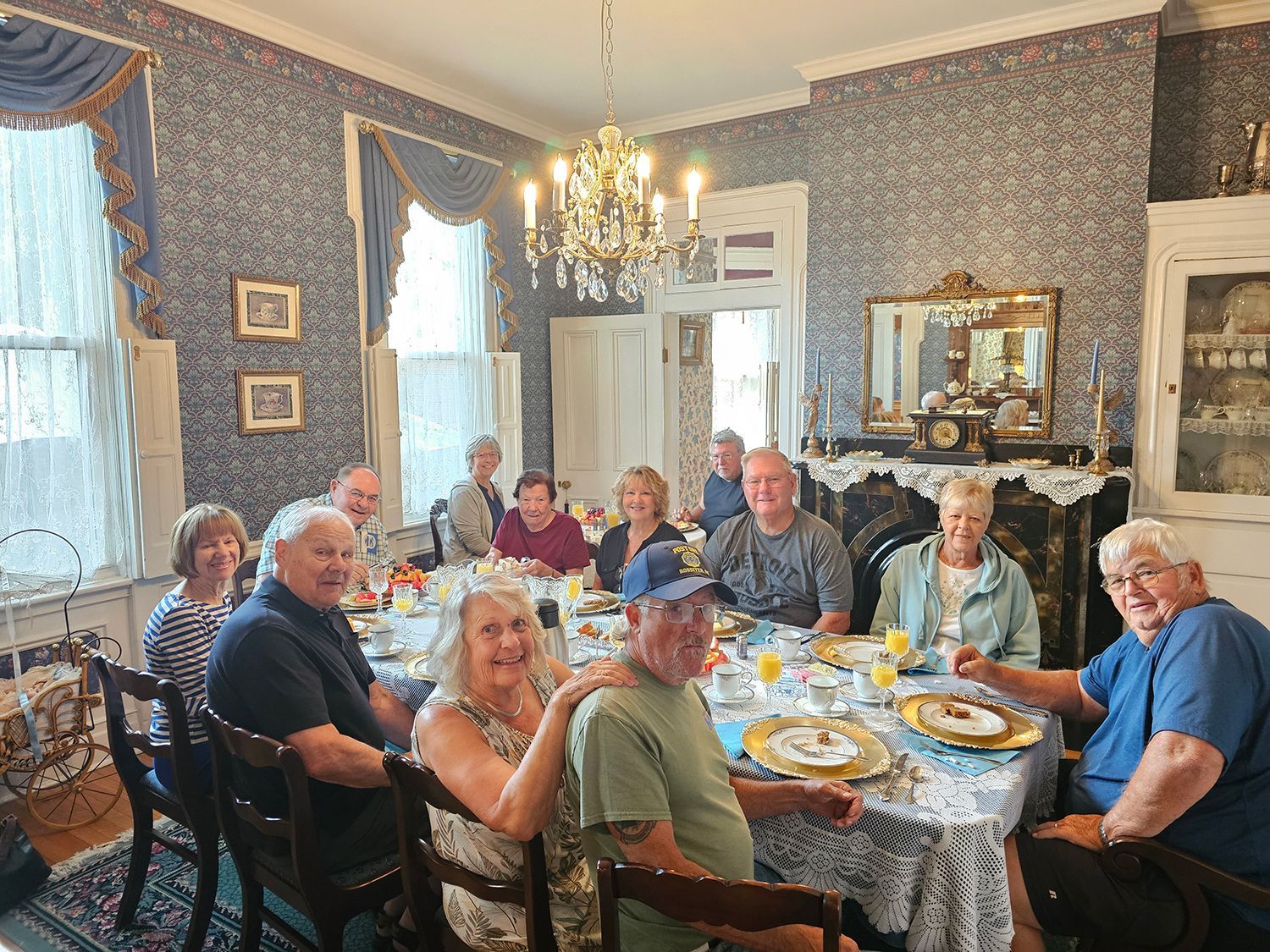 A group of people seated around a dining table, enjoying a meal in an ornate room with blue patterned walls and a chandelier.