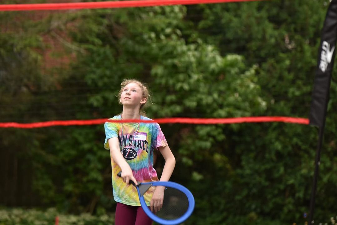 A young girl playing a game with a blue racket under a red net in a park, looking up with focused expression.