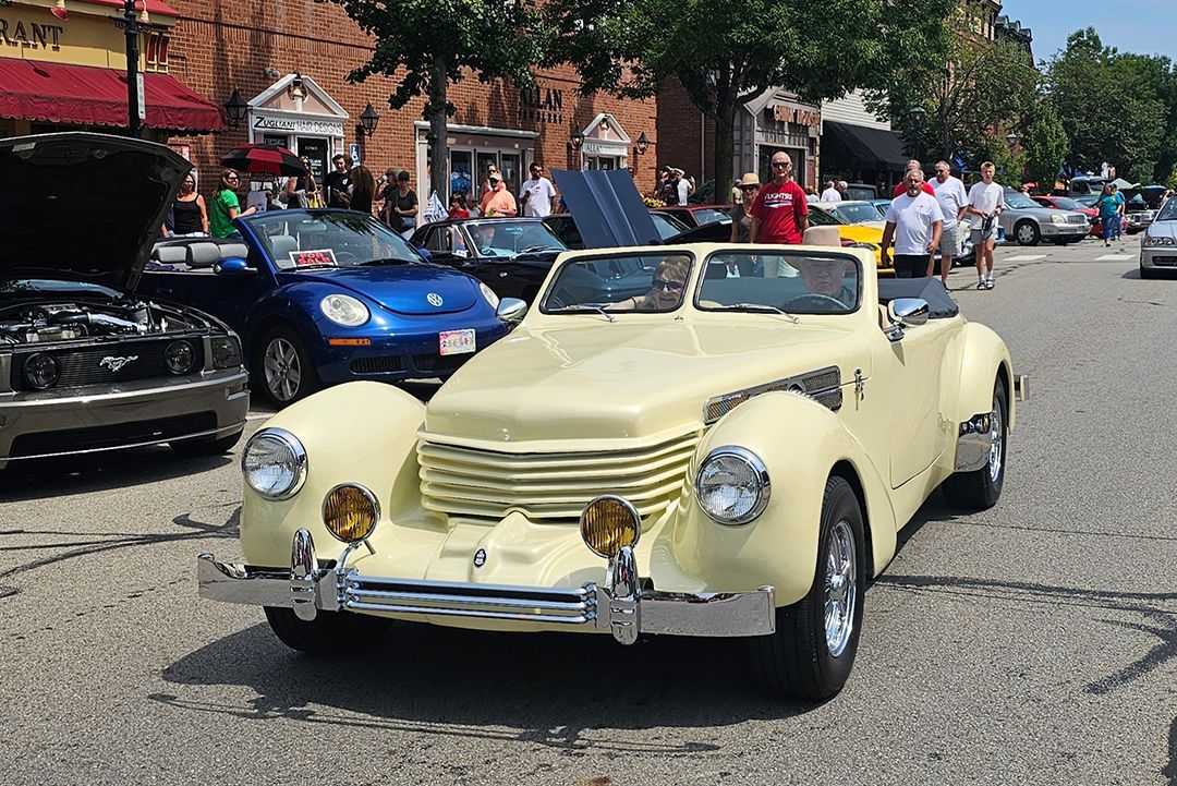 Yellow vintage car on display at an outdoor car show on a sunny day.  People are visible in the background.