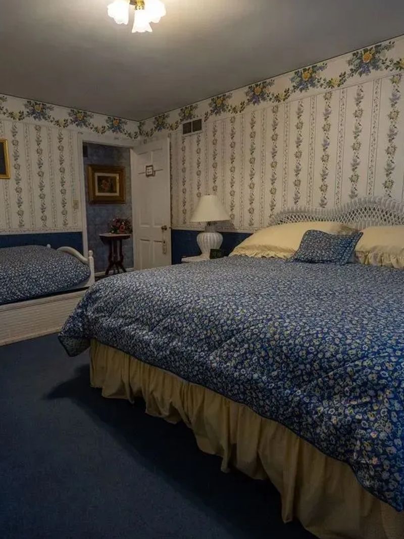 Bedroom with a large bed covered in blue floral bedding, a smaller bed, and floral wallpaper. A lamp sits on a nightstand.