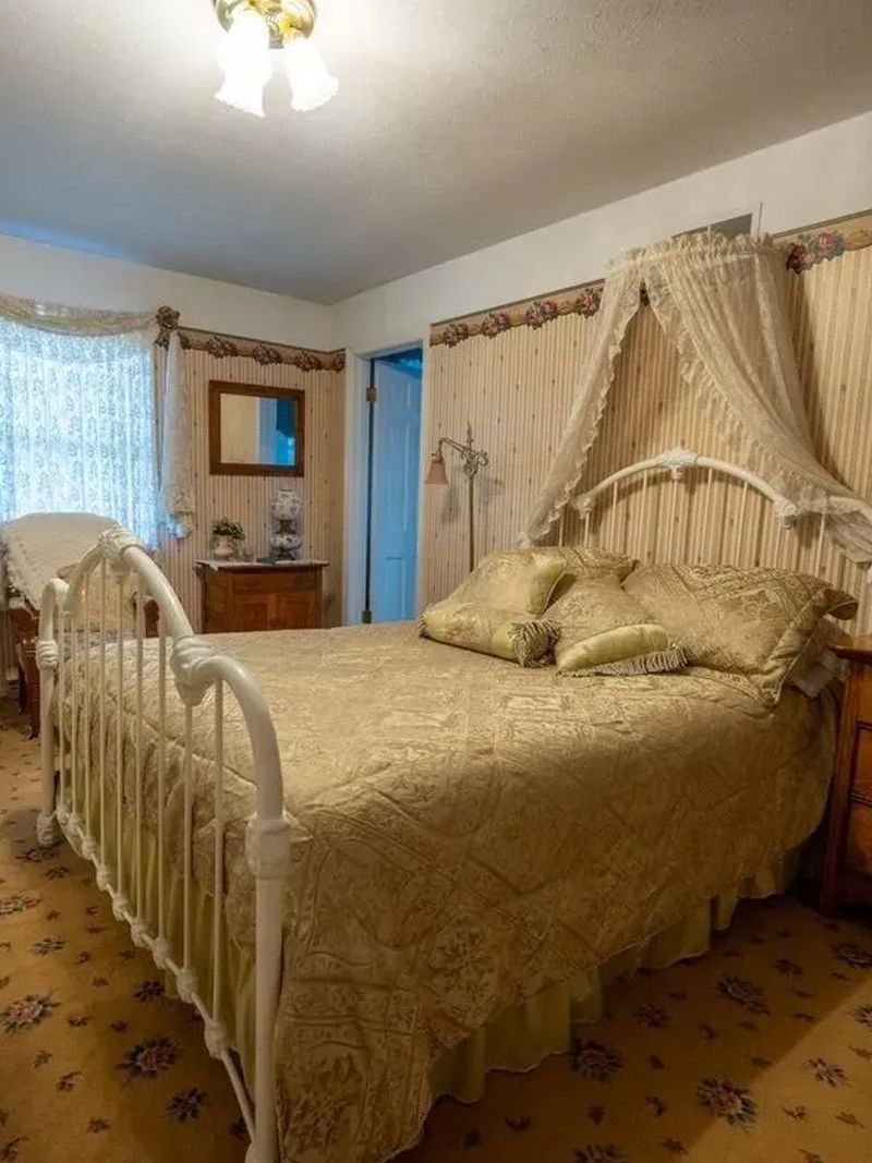 Bedroom with a white metal bed frame, gold patterned bedding, and a canopy. Beige walls, a patterned rug, and antique furniture.