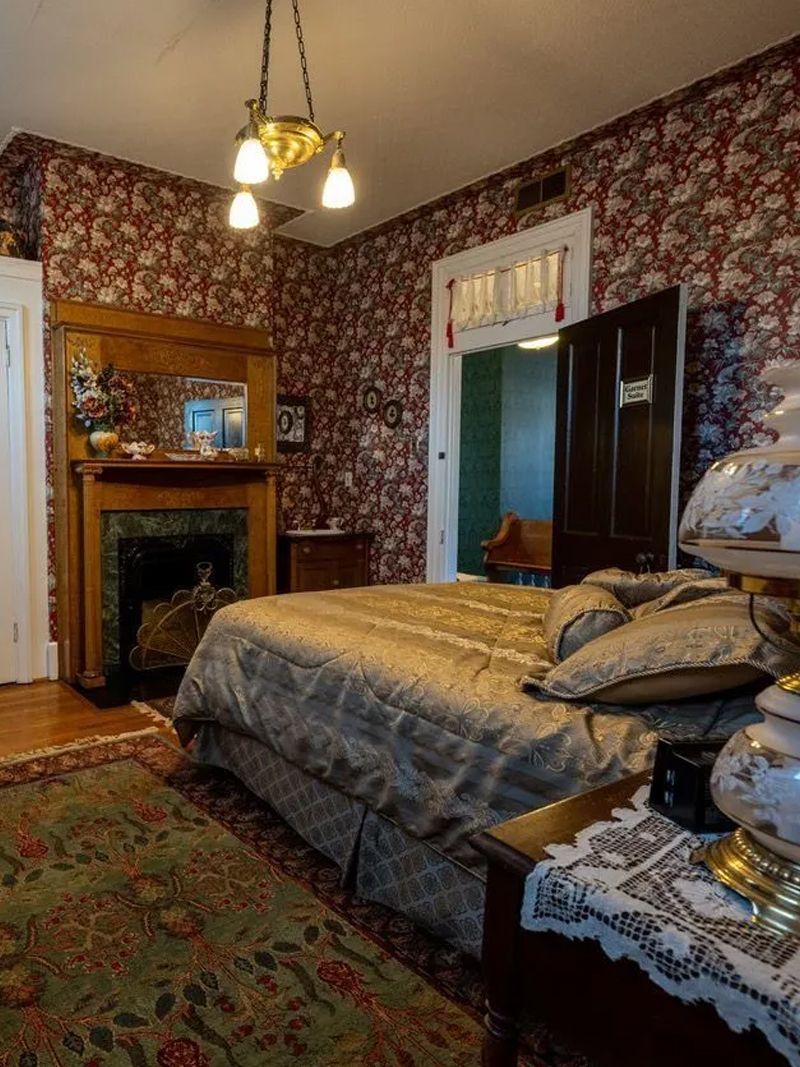 Bedroom with floral wallpaper, fireplace, antique furniture, and a bed with a patterned quilt. A doorway leads to another room.