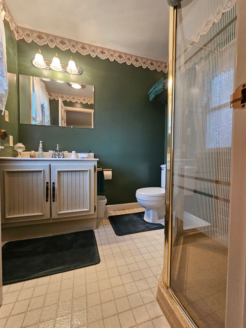 Bathroom with dark green walls, light wood vanity, and a glass shower door.  White toilet and beige tile floor.