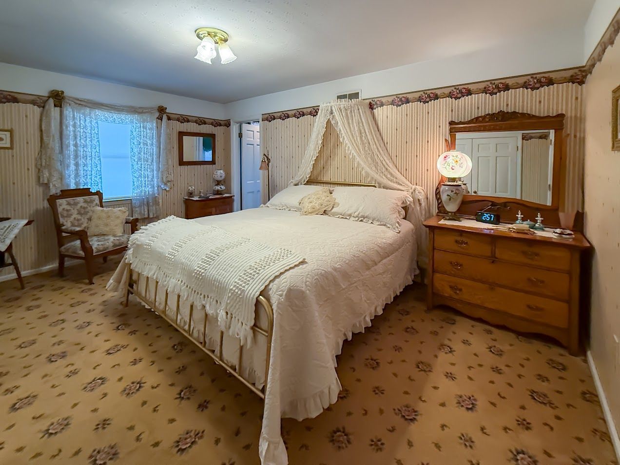 Bedroom with floral wallpaper, canopy bed, antique dresser, patterned carpet, and window with curtains.