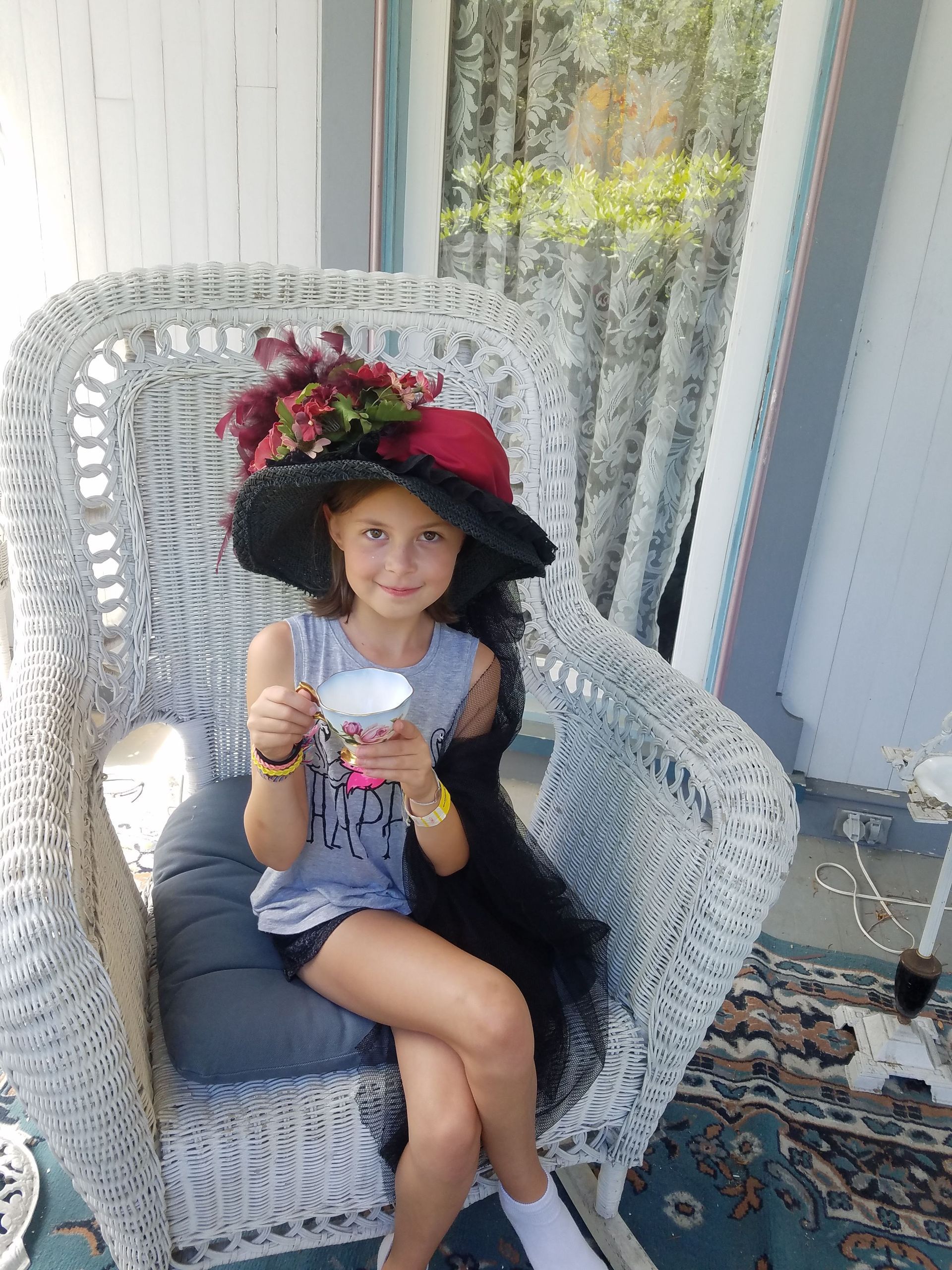 A little girl is sitting in a wicker chair holding a cup of tea.