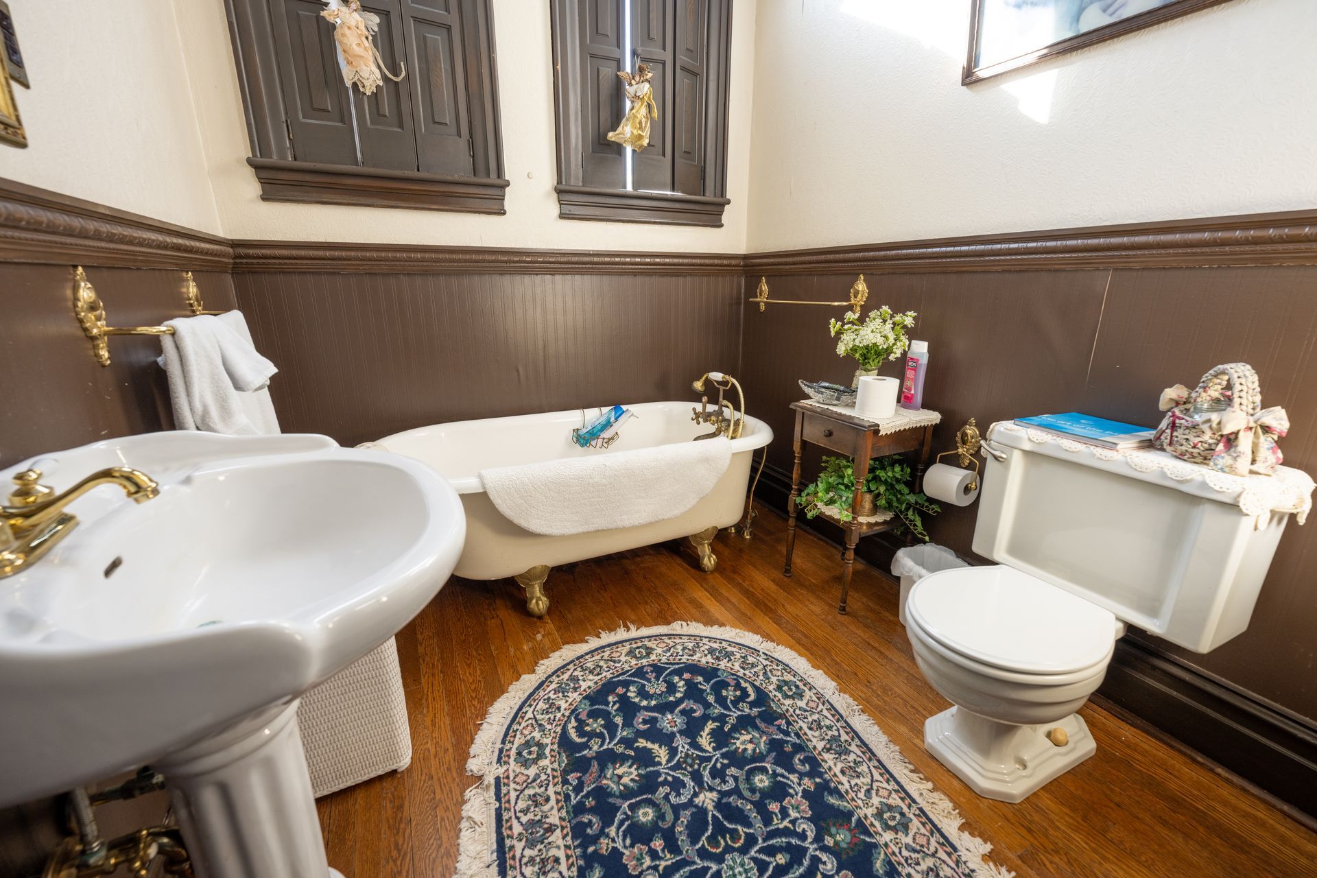Bathroom with clawfoot tub, pedestal sink, and toilet. Brown walls, gold fixtures, and small blue rug.