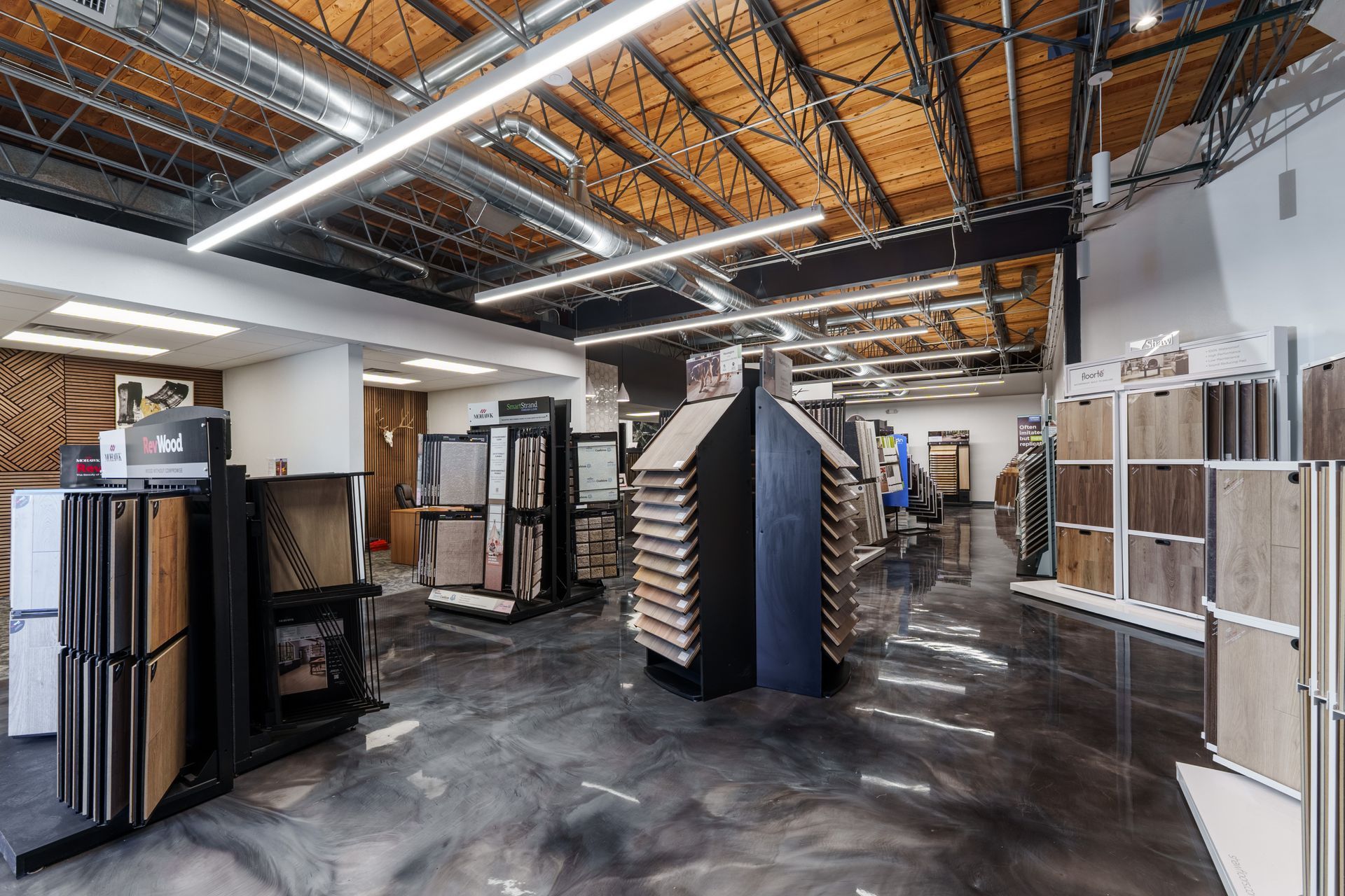 Spacious retail showroom with flooring displays, black shelving, and exposed ceiling beams under bright lights
