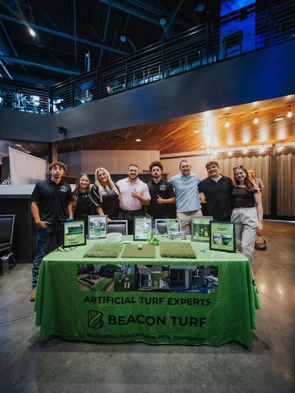 Group of people standing behind a table with artificial turf samples. Beacon Turf sign and interior setting.