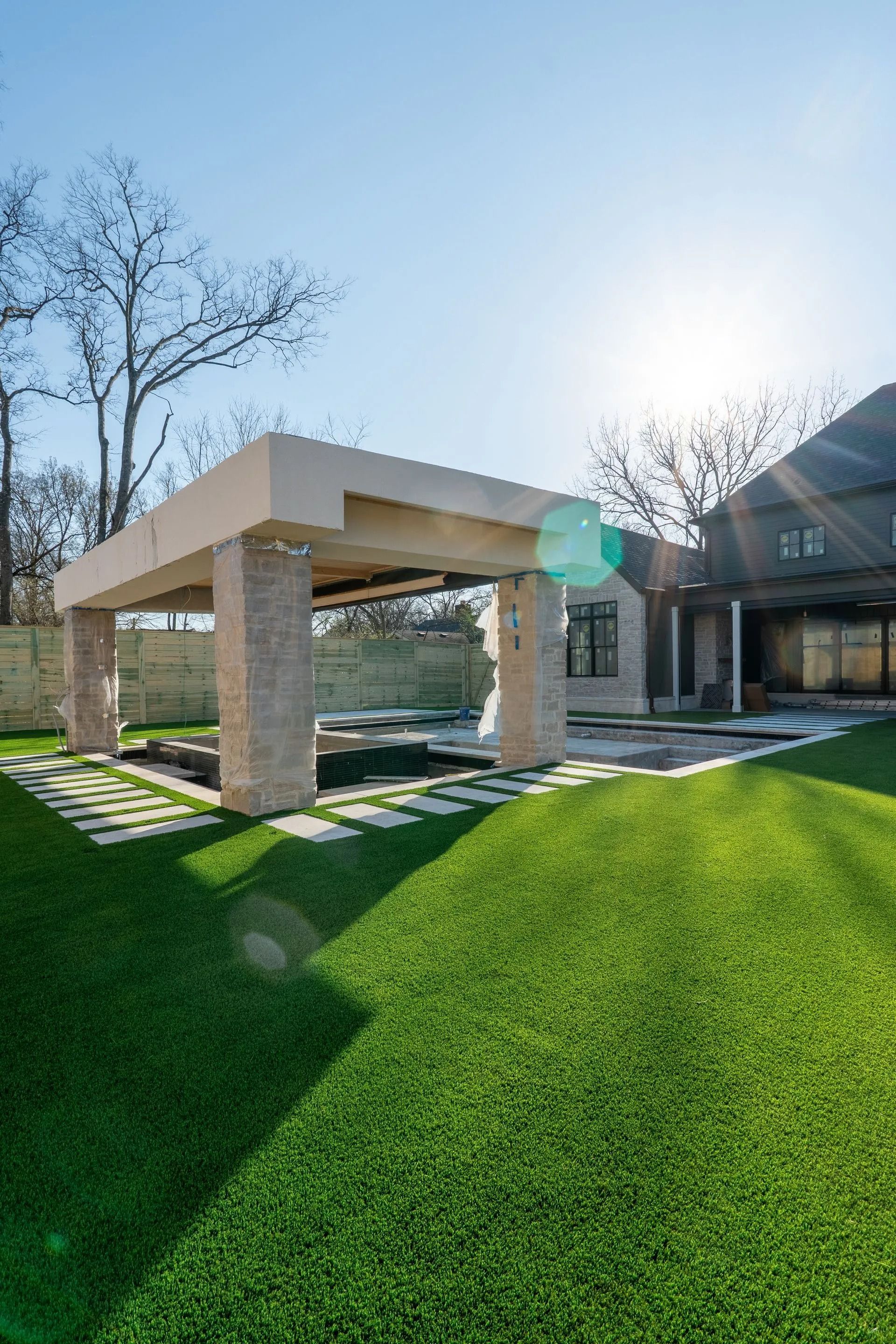 A modern outdoor pavilion with stone columns sits on a green lawn, near a house. Bright sun shines.