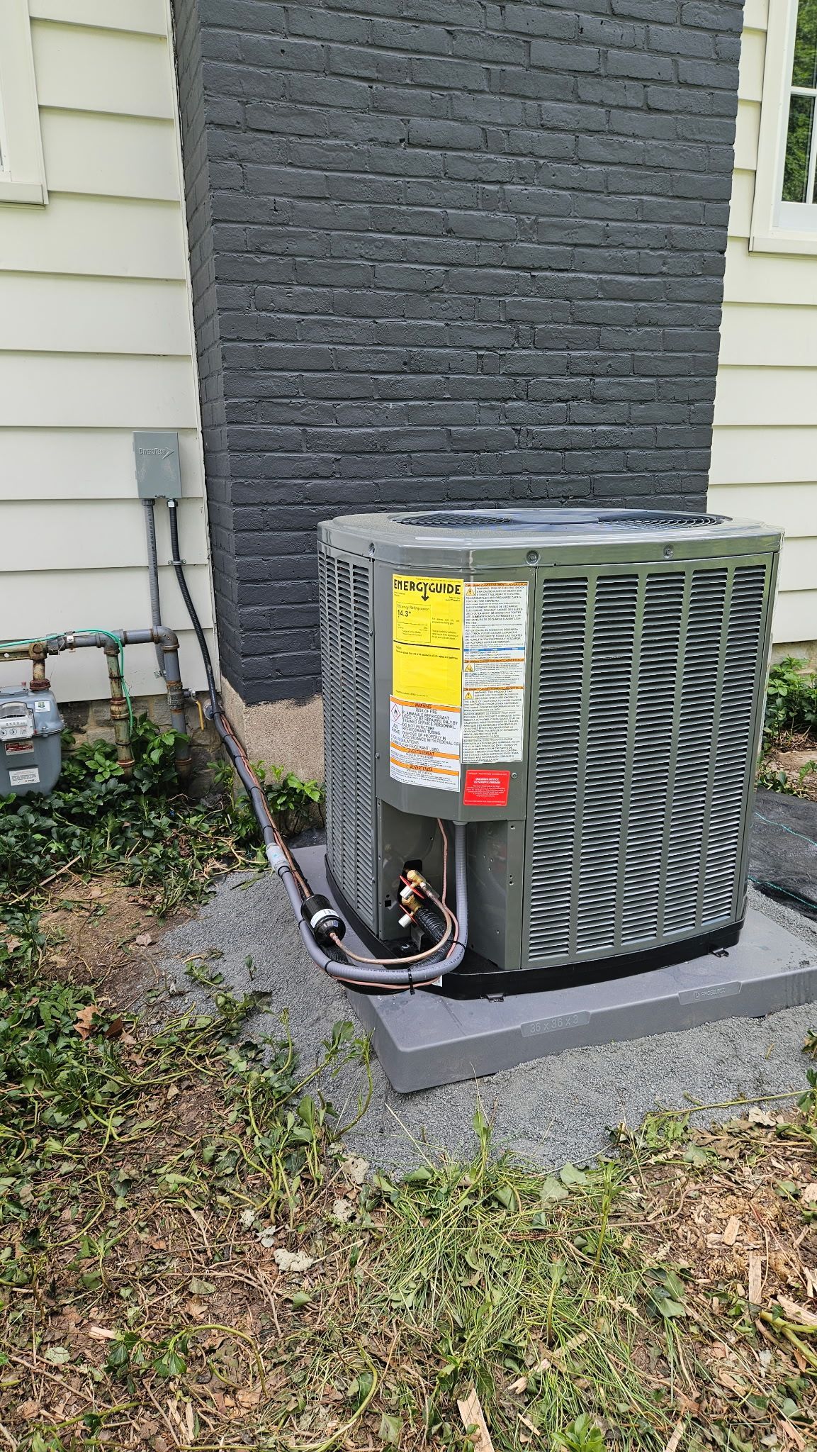 Outdoor air conditioning unit beside a house, on a gravel pad near siding and grass.