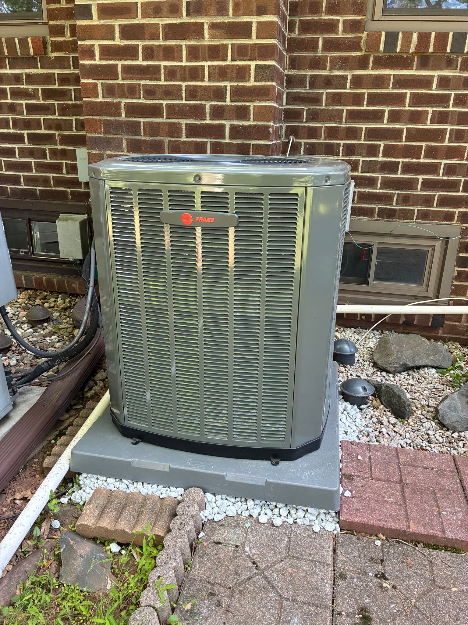 Outdoor HVAC condenser unit beside a brick house wall, set on a concrete pad with landscaping stones nearby