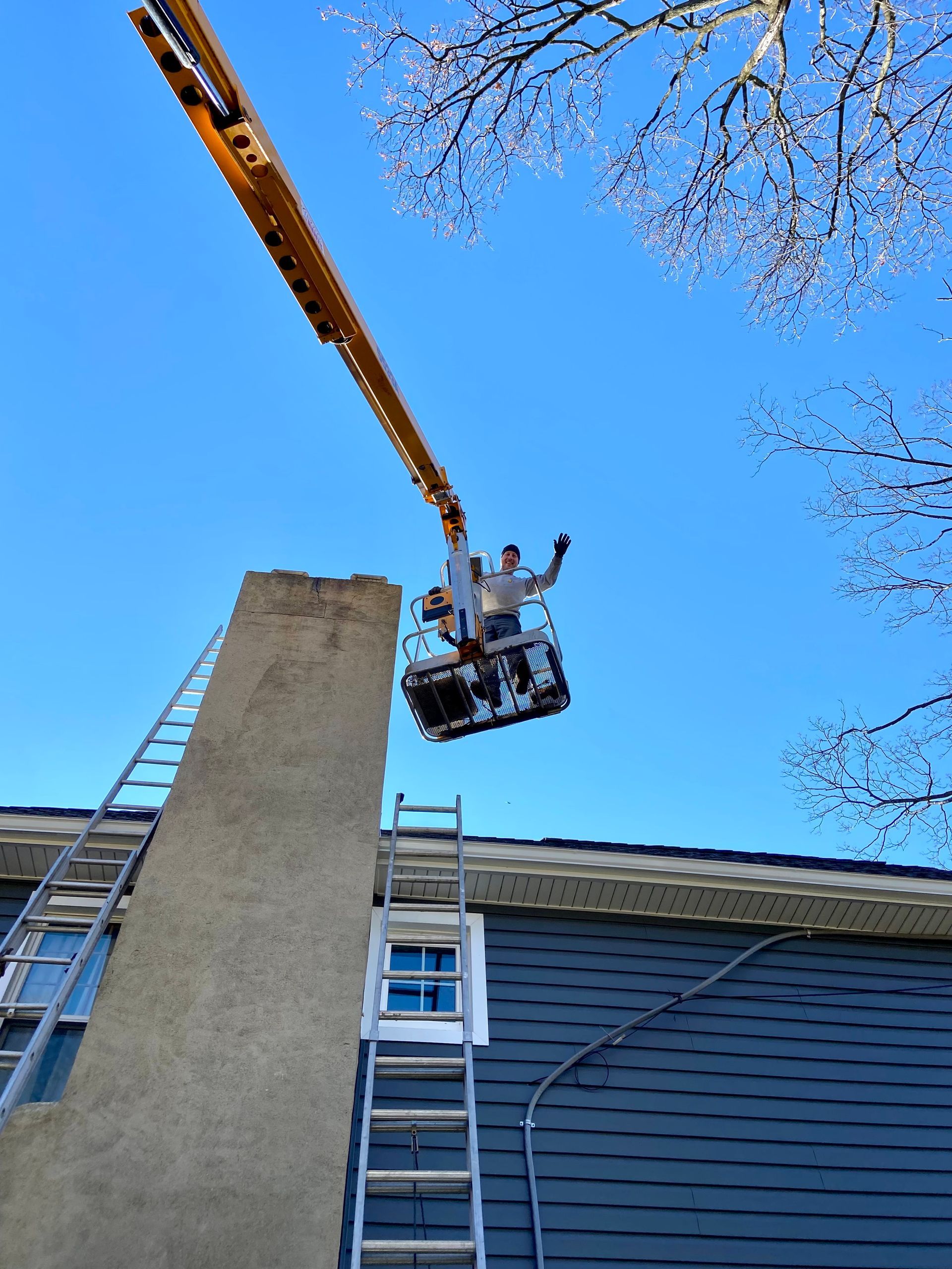 Two workers in a cherry picker beside a building roof, reaching up near a tall brick chimney.
