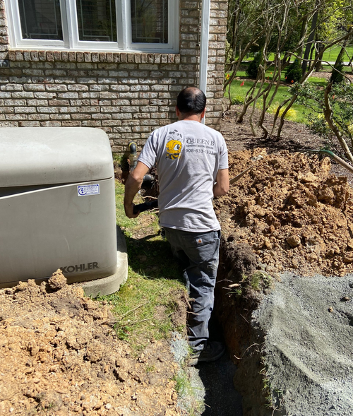 Worker in gray shirt stands in a narrow trench beside a brick house and dirt pile.