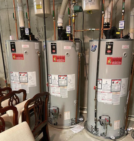 Three gray water heaters in a utility room with pipes and valves overhead