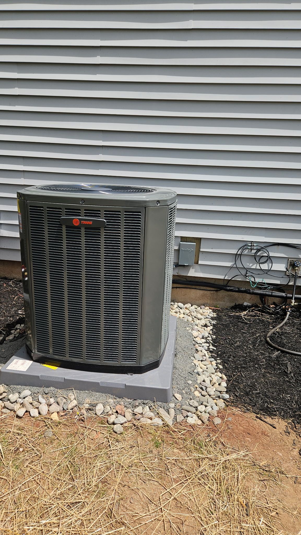 Outdoor air conditioner unit beside a house with gray siding and gravel ground