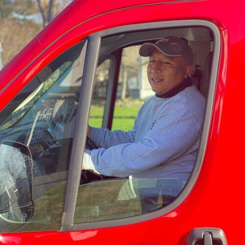 Man sitting in the driver’s seat of a red vehicle, wearing a cap and light shirt, looking out the window.
