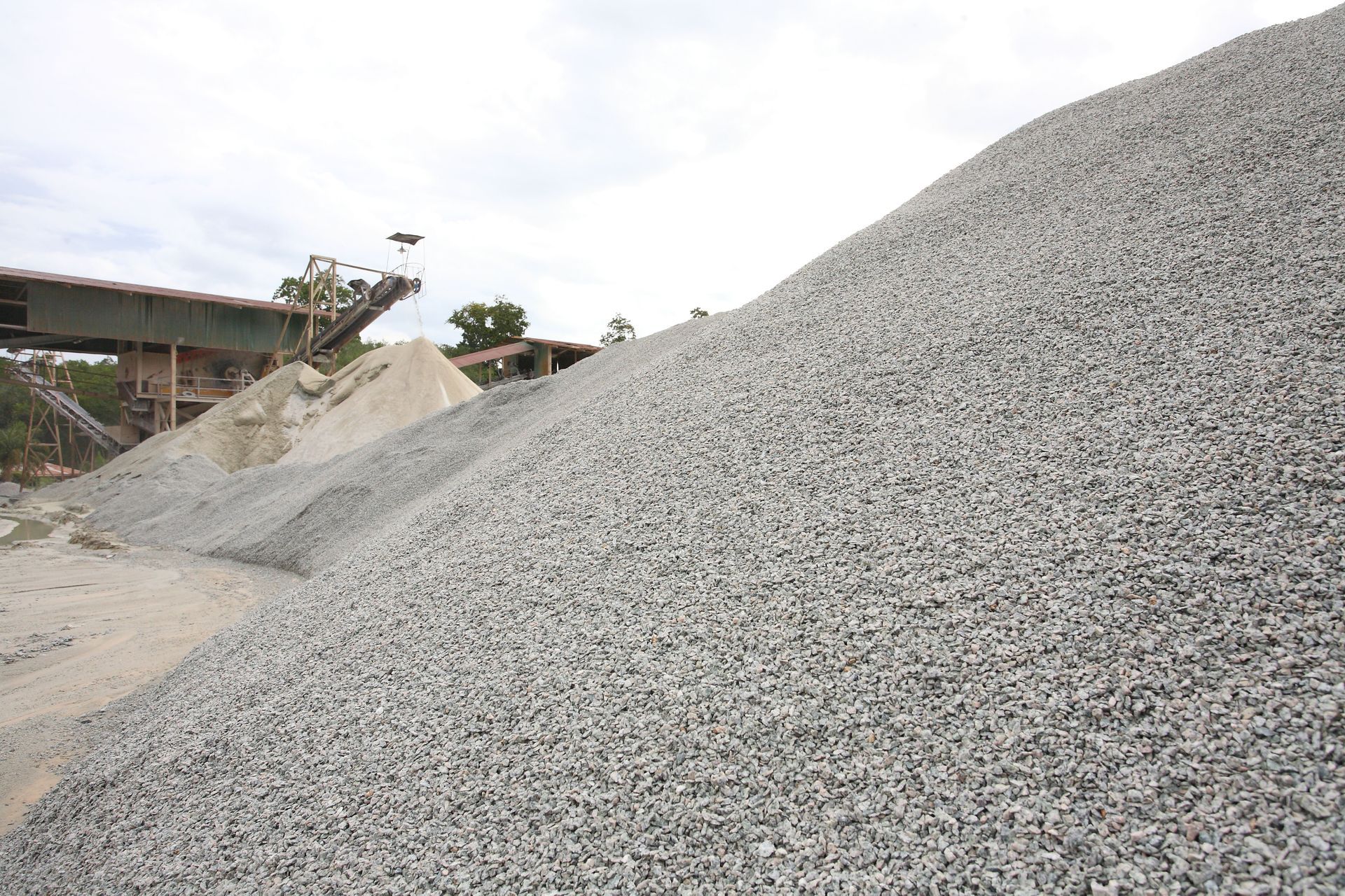 Pile of gray gravel in a quarry, with machinery in the background under a cloudy sky.