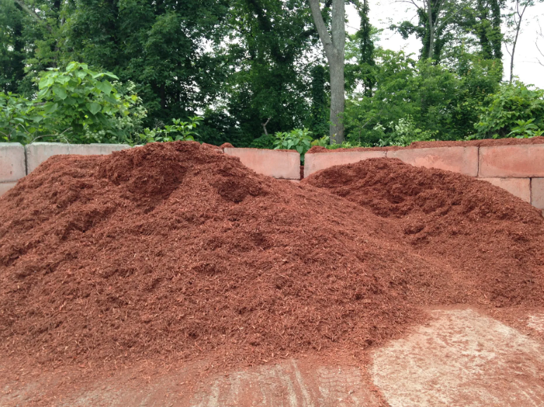 Piles of reddish-brown mulch against a brick wall, with greenery in the background.