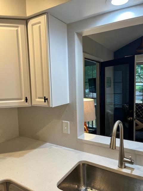 Kitchen sink and white cabinets beside a window looking into a dark living area