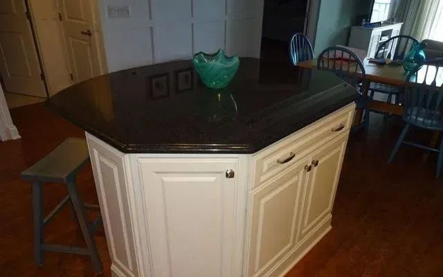 Kitchen island with dark countertop, white cabinets, bar stool, and dining table in background