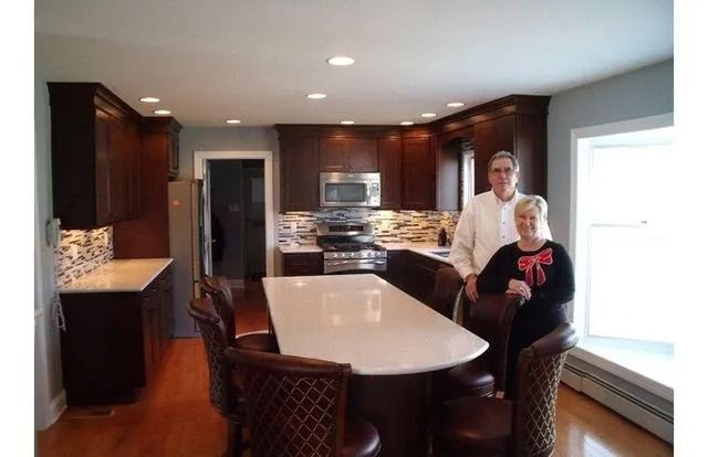 Modern kitchen with two people standing by a white island, dark cabinets, and large windows.