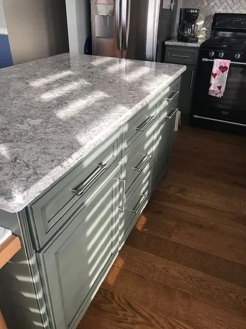 Kitchen island with light gray granite countertop and sage green cabinets beside a stove.