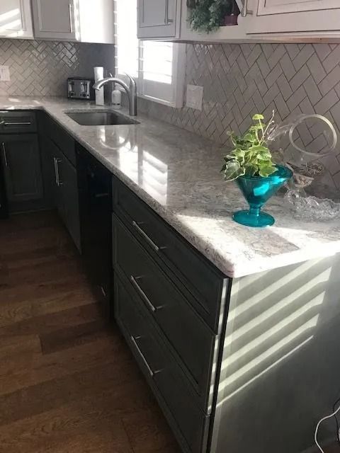 Modern kitchen with dark cabinets, gray herringbone backsplash, and sunlight on a white countertop with a small plant.