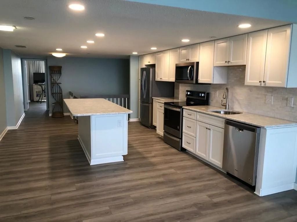 Modern open-concept kitchen with white cabinets, gray island, stainless appliances, and wood-look floors.