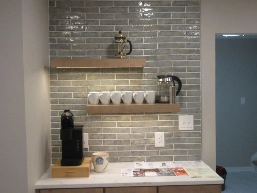 Coffee station on a tiled wall shelf with a black machine, cups, kettle, and magazines on a counter