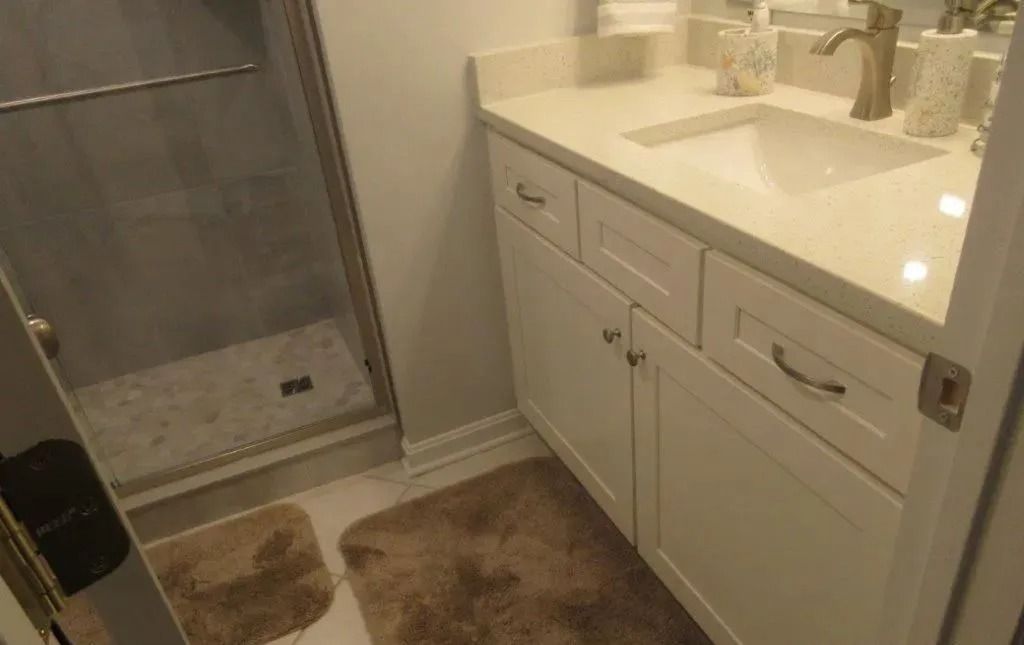 Bathroom vanity with white cabinets, double sink, mirror, shower stall, and bath mats on tile floor