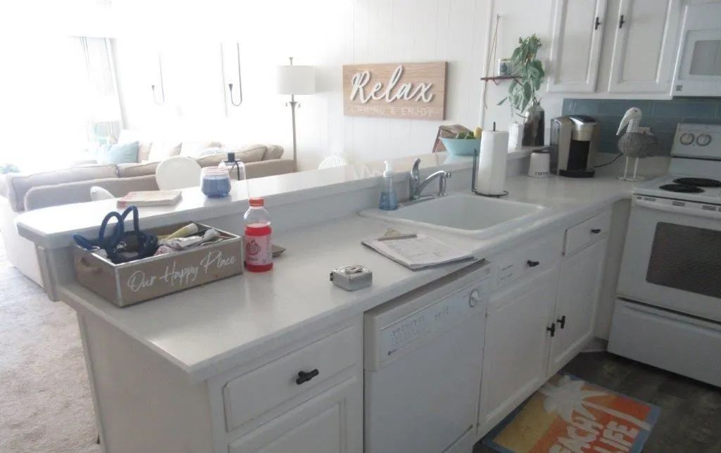 Bright white kitchen counter with sink, dish rack, soap, and decorative sign by a sunny window