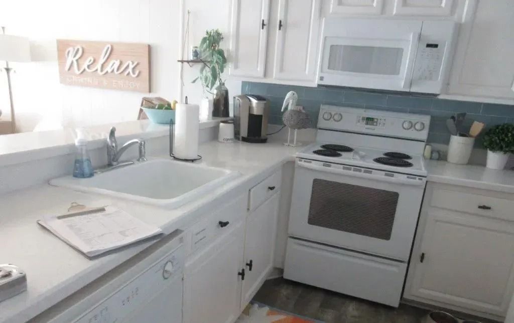 Bright white kitchen with stove, sink, and countertop decor, including a “Relax” sign by the window