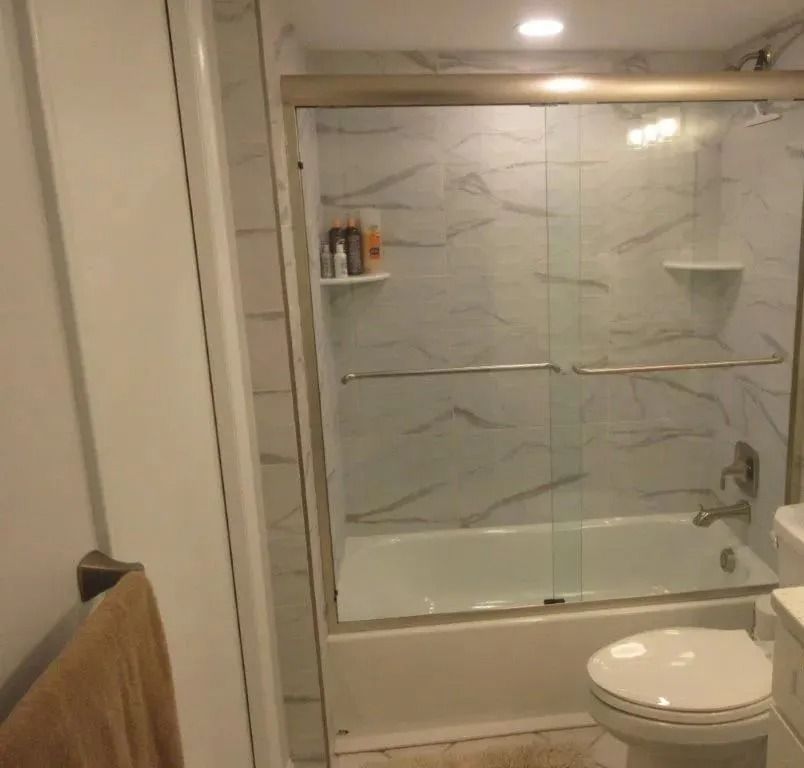 Bathroom with glass shower/tub combo, marble-look tile, towel bar, and toilet in foreground