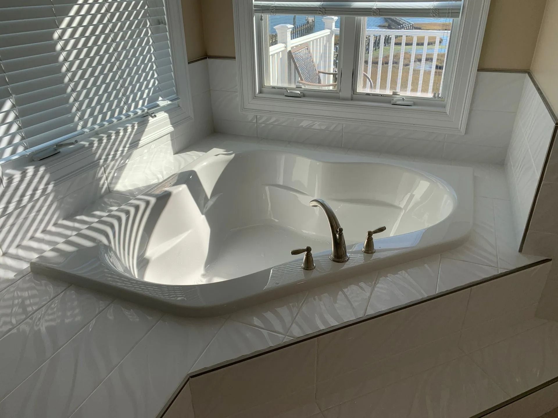 Corner bathtub beside a window with sunlight and blinds casting striped shadows on white tile.