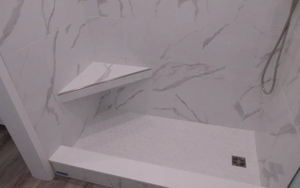 Empty shower stall with white marble-look walls, a shelf, and a built-in bench.
