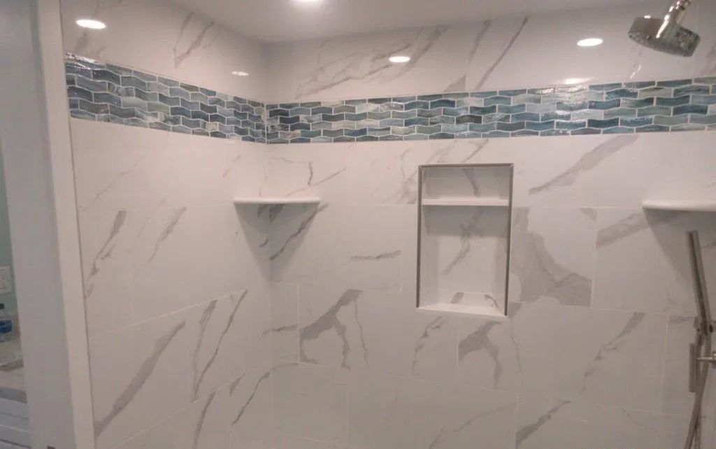Modern tiled shower with marble-look walls, blue mosaic border, recessed shelves, and overhead lights