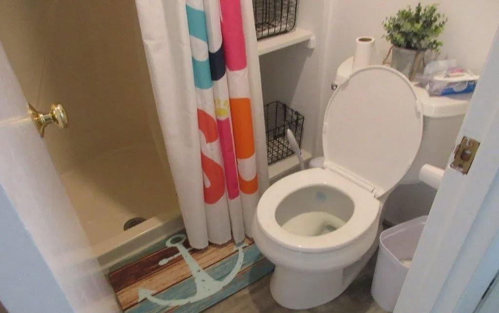 Small bathroom with open toilet, colorful shower curtain, and bath mat beside a sink.