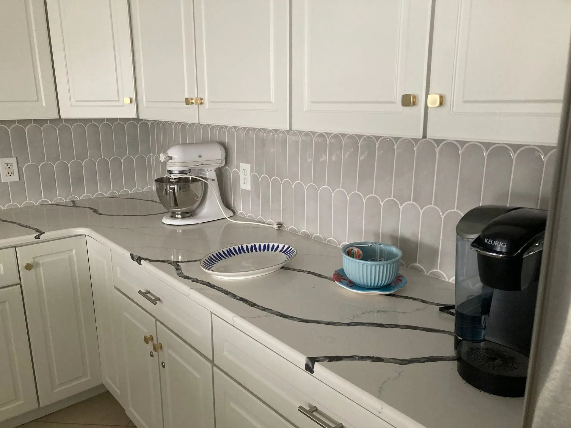 White kitchen countertop with a stand mixer, platter, mug, and coffee maker under white cabinets