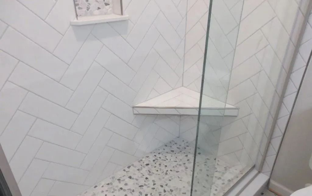 White-tiled shower with glass door and corner shelf, featuring small patterned floor tiles.