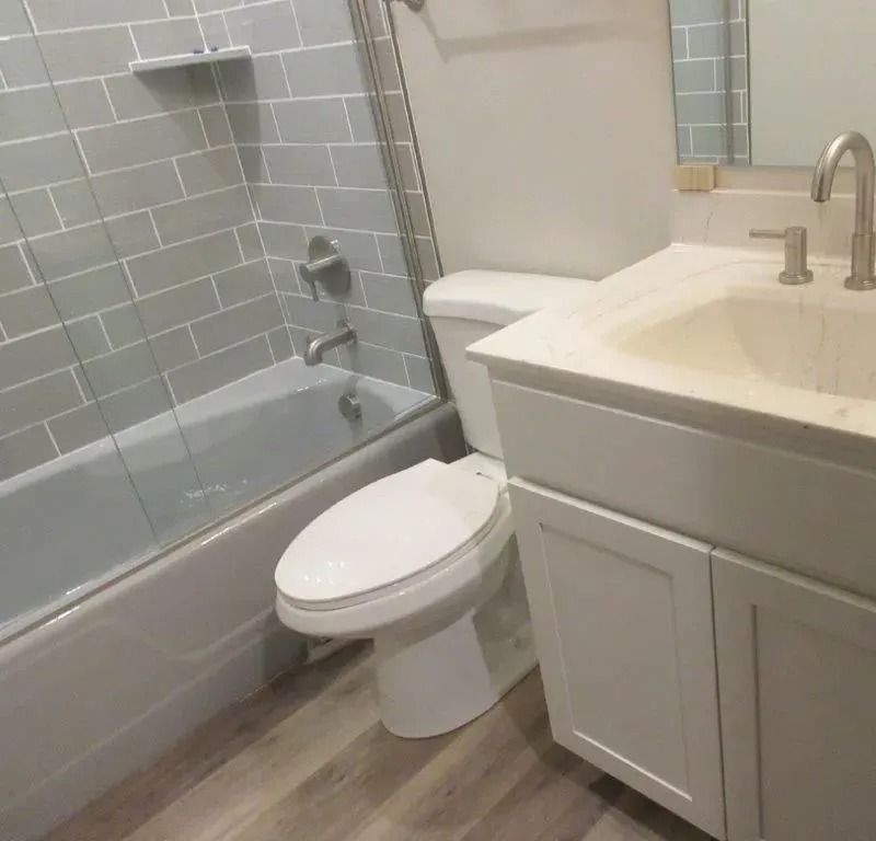 Small bathroom with white vanity, toilet beside a bathtub, and gray tile walls.