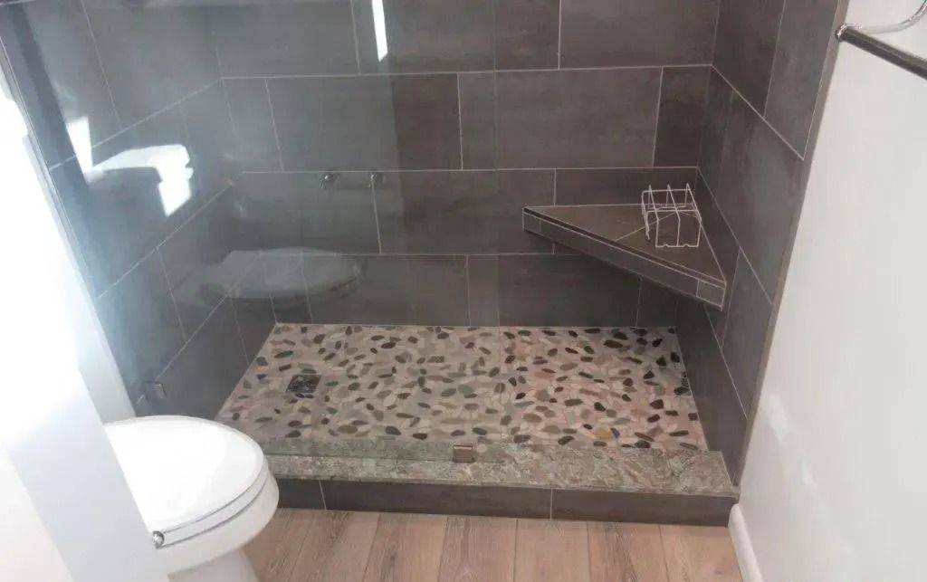 Small tiled walk-in shower with pebble floor, built-in corner shelf, and partial toilet view.