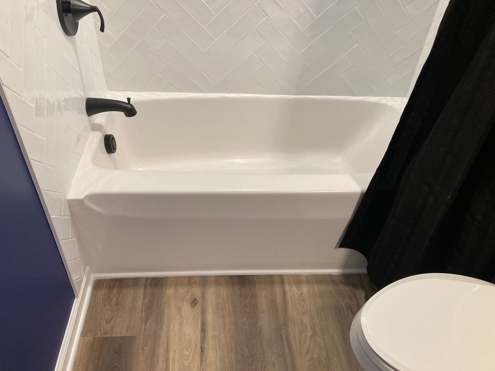 Small bathroom with white tub, black fixtures, wood-look floor, and a partial toilet in the foreground