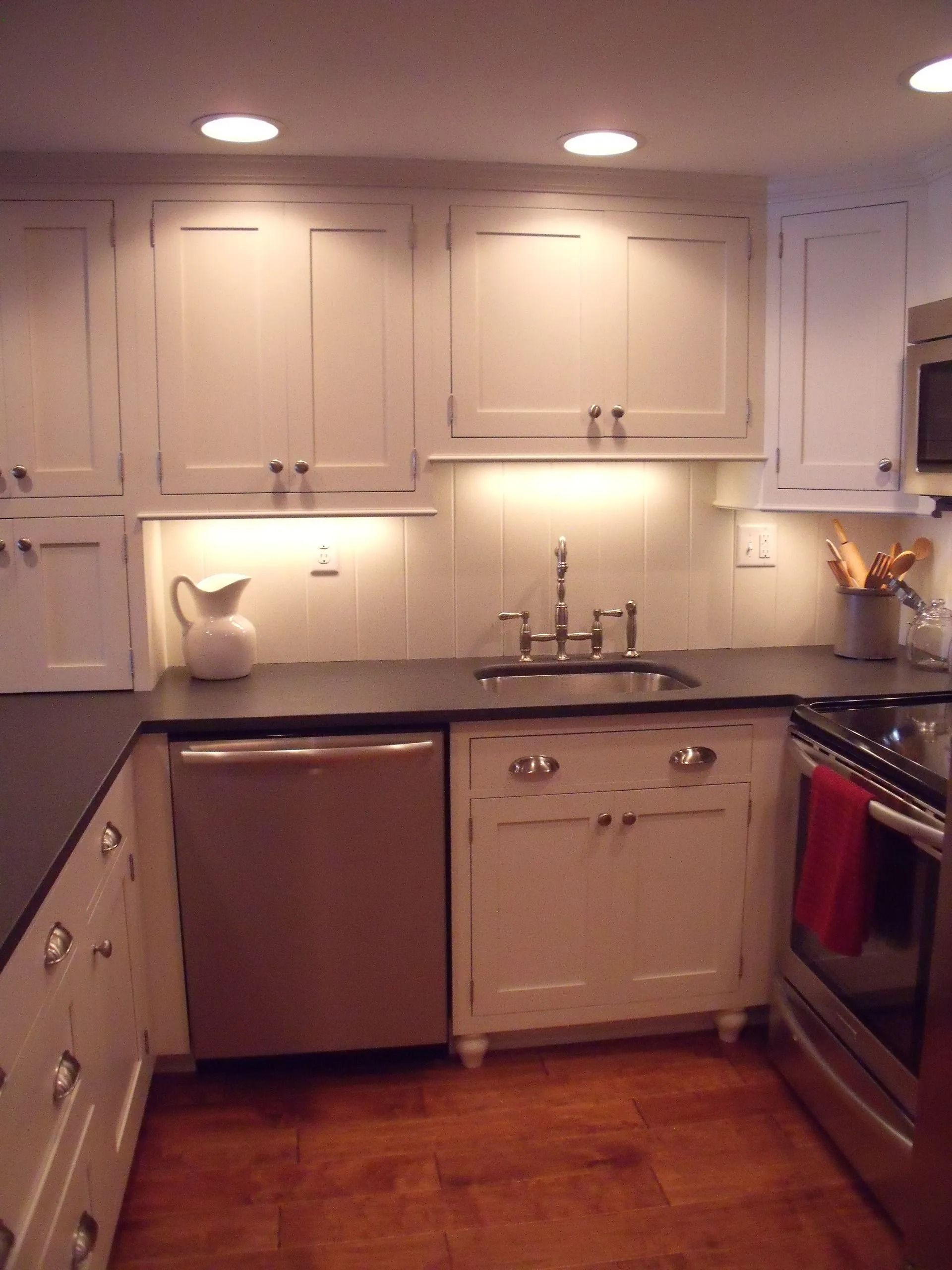 Small white kitchen with wood floors, white cabinets, stainless appliances, and under-cabinet lighting.
