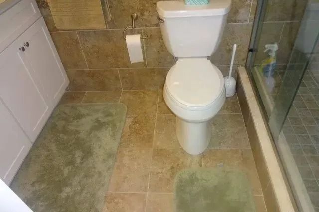 Small bathroom with toilet, beige tile floor, green bath mats, and a glass shower enclosure.