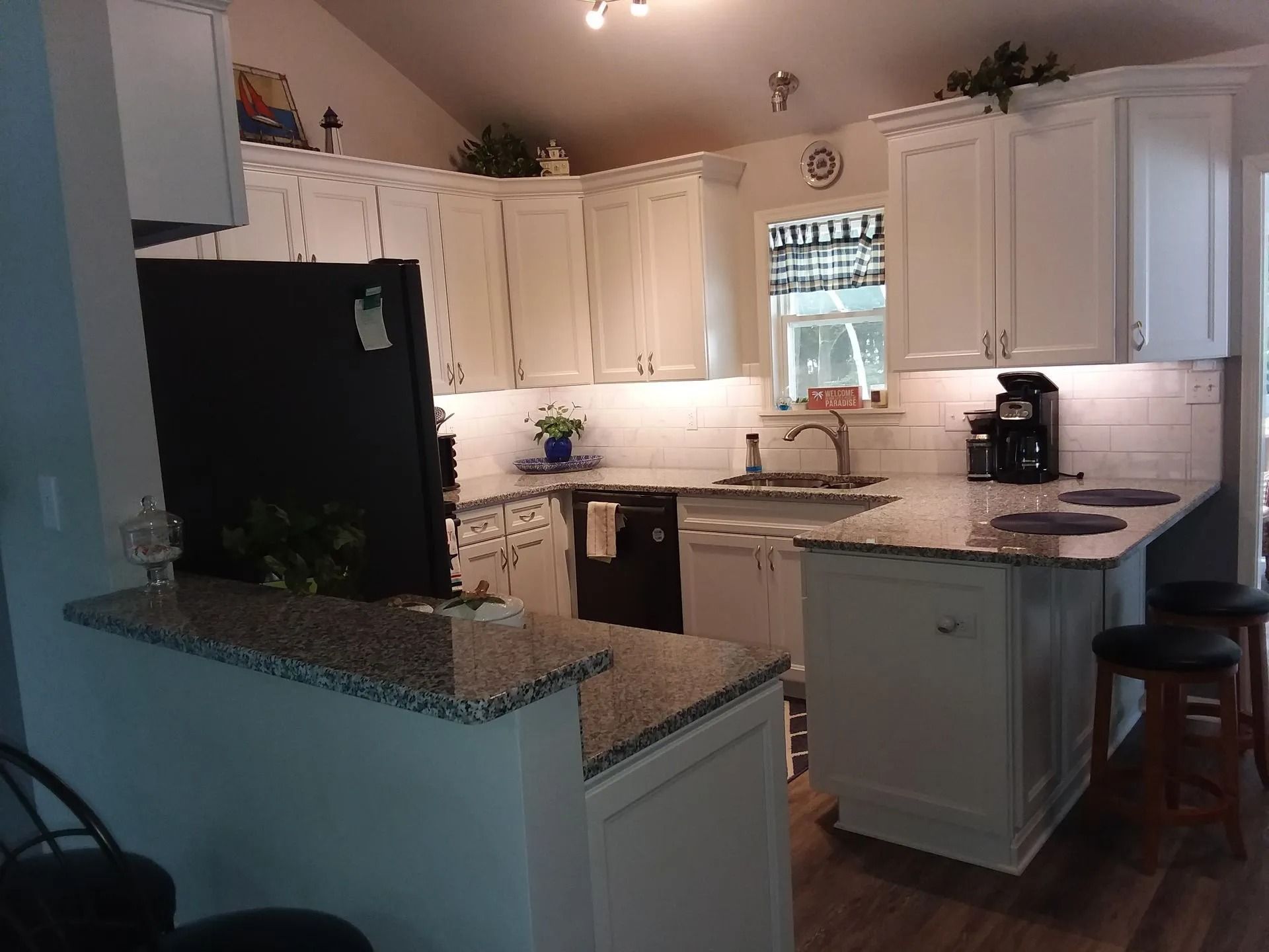 Bright kitchen with white cabinets, black fridge, granite island, and coffee maker near a window.