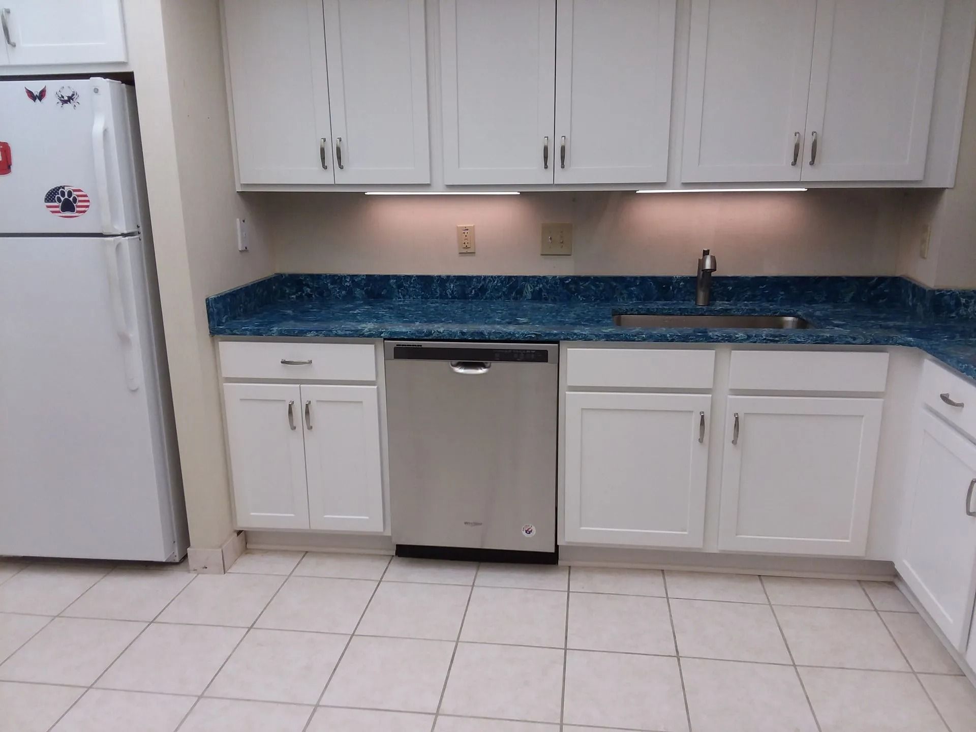 White kitchen with blue countertop, white cabinets, stainless dishwasher, and tiled floor.