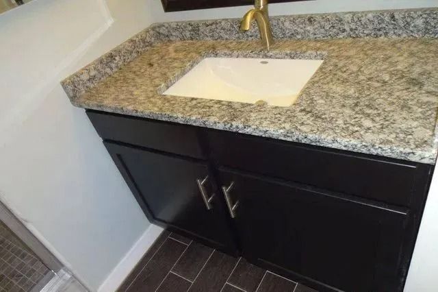 Black bathroom vanity with granite countertop, white sink, and gold faucet on dark tile floor
