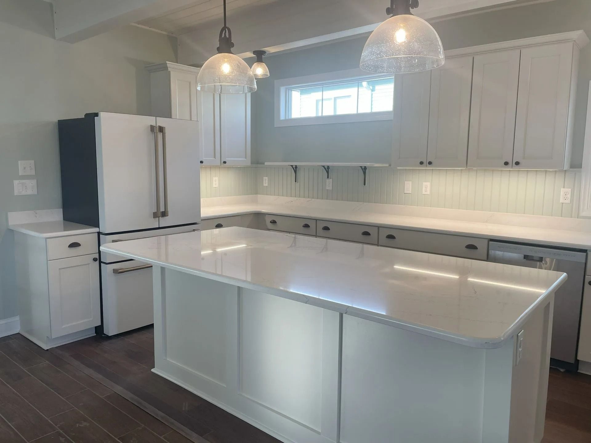 Bright white kitchen with island, pendant lights, stainless fridge, and dark wood floors.